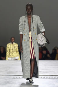 Model walking a fashion runway in a long houndstooth coat, open at the front, paired with a burgundy and beige striped skirt and a pale embellished bag swung on the right arm.