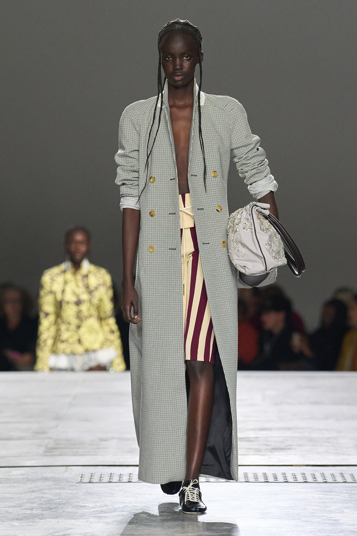 Model walking a fashion runway in a long houndstooth coat, open at the front, paired with a burgundy and beige striped skirt and a pale embellished bag swung on the right arm.