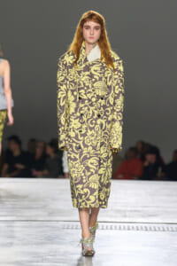Model walking the runway in a long brown coat with a large yellow floral pattern and matching yellow-green pants; the model wears jeweled sandals.
