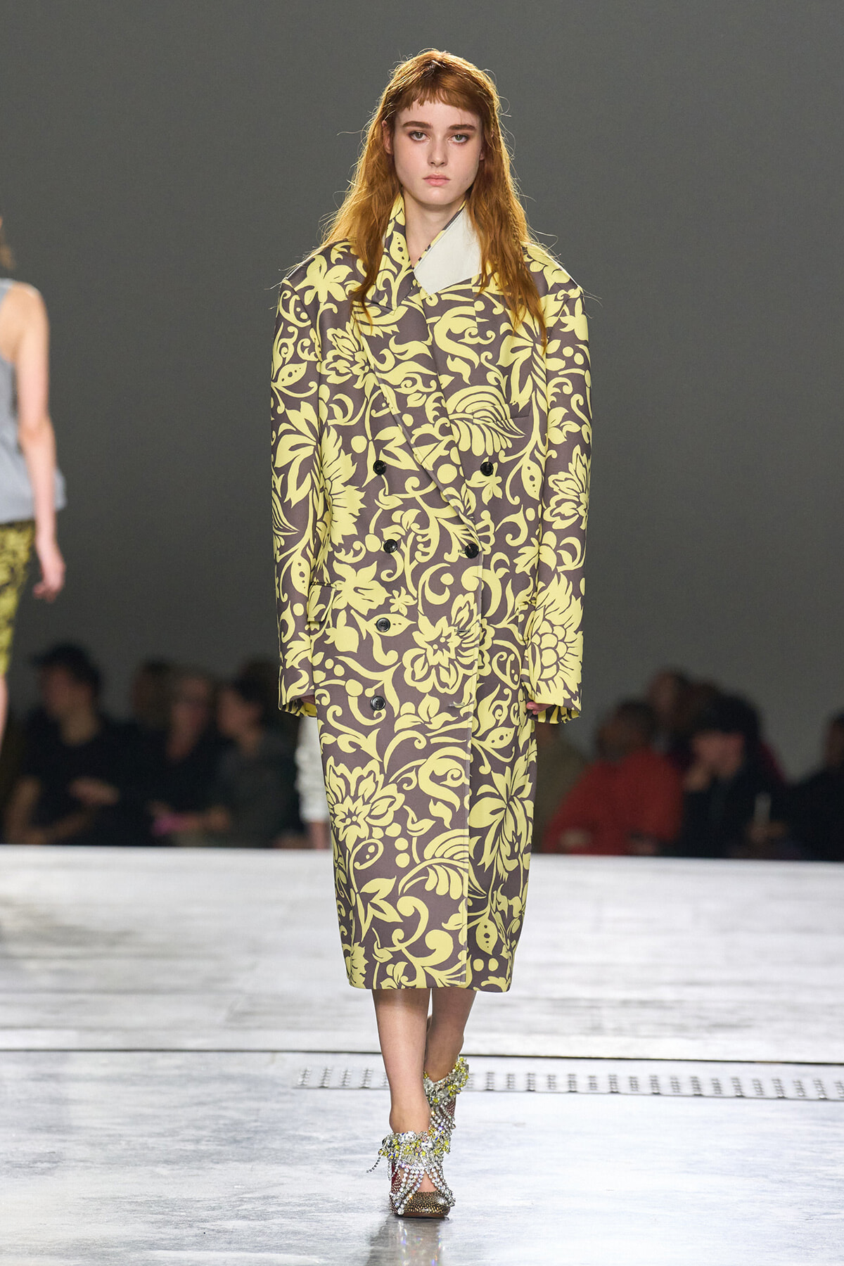 Model walking the runway in a long brown coat with a large yellow floral pattern and matching yellow-green pants; the model wears jeweled sandals.