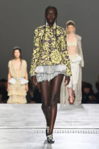 Front view of a dark-skinned model walking a runway in a bold olive-yellow floral print blazer and a gray layered ruffle mini-skirt, with black lace-up shoes.