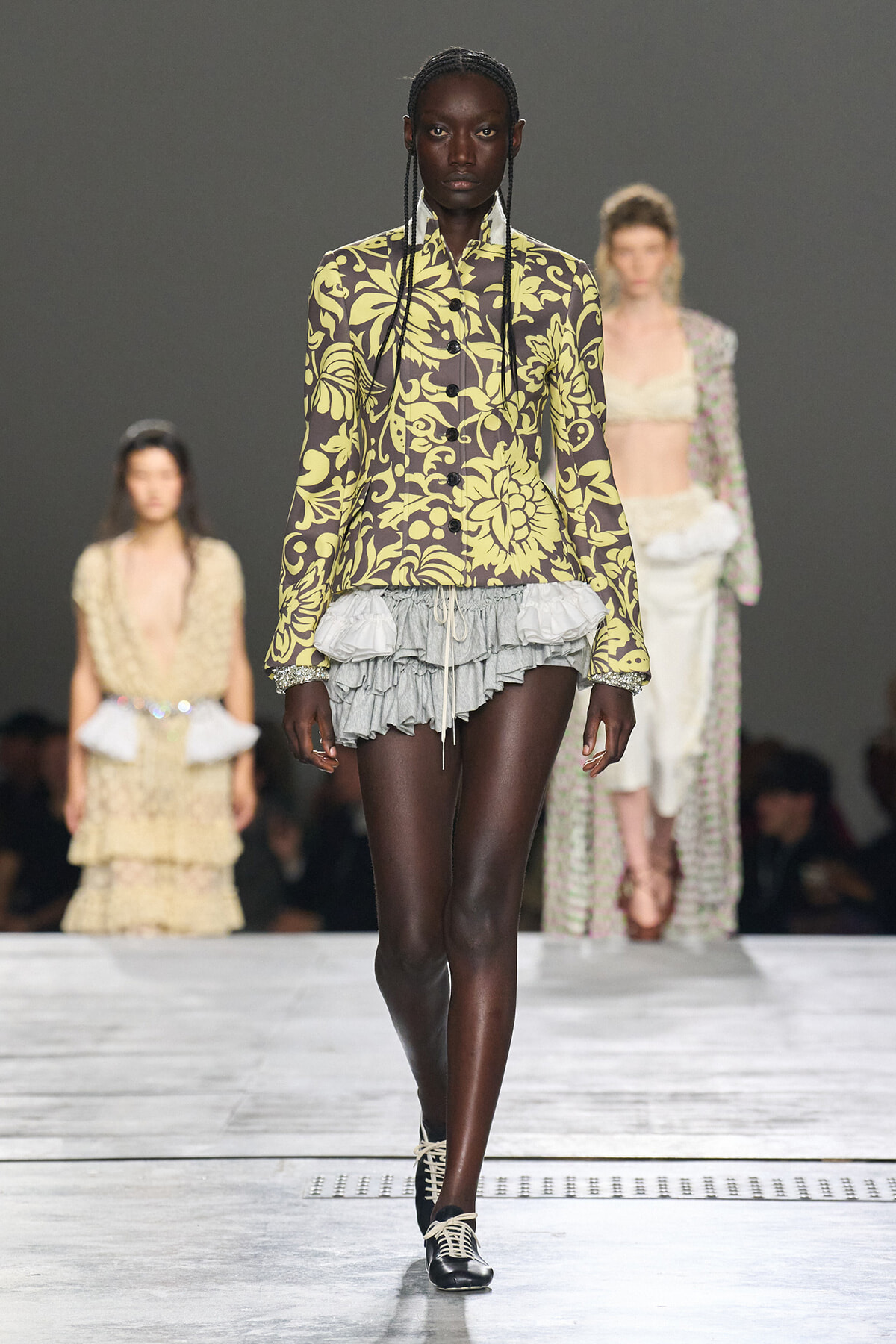 Front view of a dark-skinned model walking a runway in a bold olive-yellow floral print blazer and a gray layered ruffle mini-skirt, with black lace-up shoes.