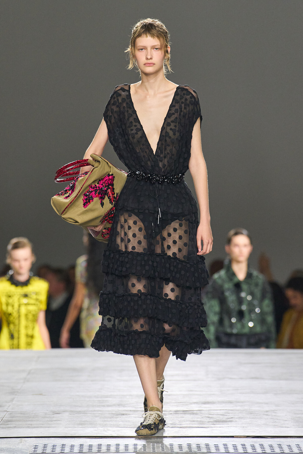 Model walking a fashion show in a sheer black polka-dot dress with tiered ruffles and a deep V neckline, carrying an embroidered beige tote bag.