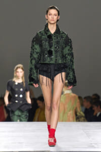 Model walks the runway in a dark green embellished jacket with black embroidery, paired with black fringed shorts and red socks shoes combo.