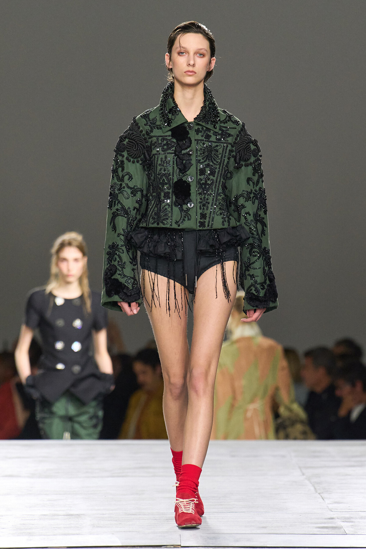 Model walks the runway in a dark green embellished jacket with black embroidery, paired with black fringed shorts and red socks shoes combo.