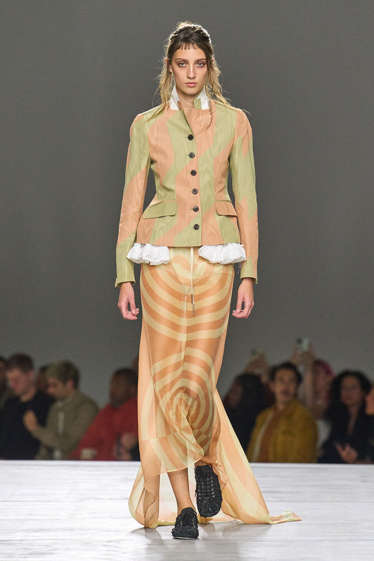 Model on runway wearing a pastel green and peach striped tailored jacket with buttons and peplum, paired with a long peach spiral-patterned skirt and black boots.
