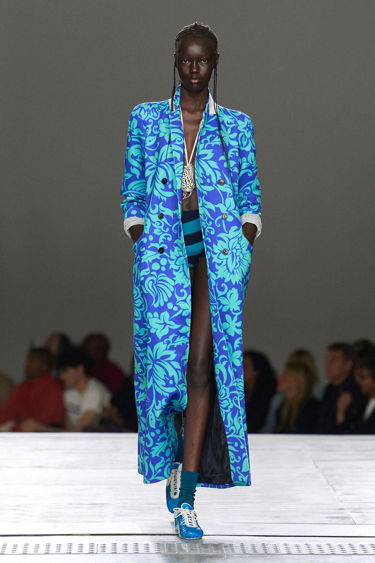 Model walks the runway in a long cobalt-blue coat with teal tropical pattern, open to reveal teal swimwear and a statement necklace.