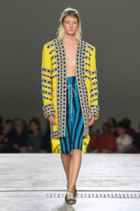 Model walking a fashion show in a bright yellow coat with blue beaded trim over teal and navy striped shorts.