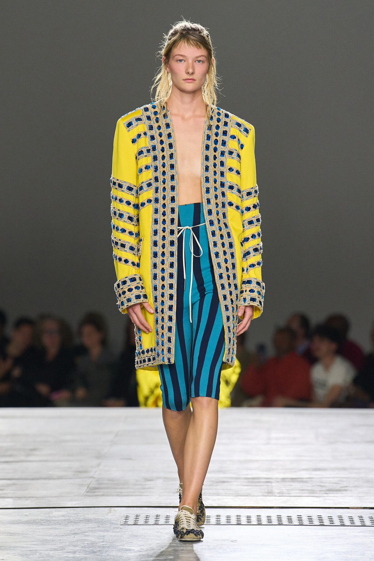 Model walking a fashion show in a bright yellow coat with blue beaded trim over teal and navy striped shorts.