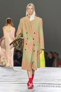 Runway model wearing a long pastel geometric trench coat, holding a patterned handbag.