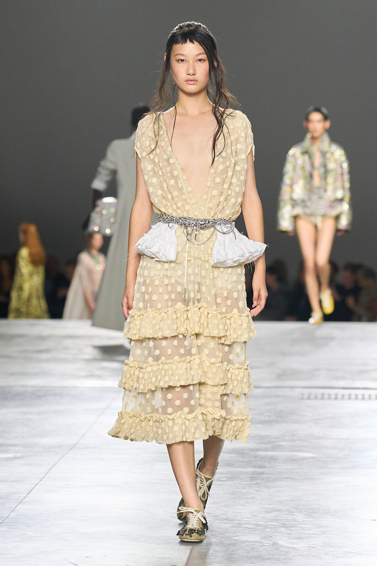 Model walking the runway in a sheer beige polka-dot dress with deep V-neck, tiered ruffles, and a sparkling belt.