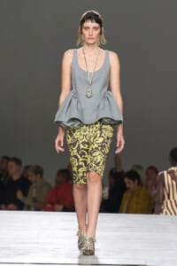 Model walking a runway in a light gray peplum top, yellow-black patterned pencil skirt, jeweled heels, and long pendant necklace.