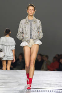 Model walks the runway in a light gray embellished bomber jacket with crystal embroidery, a gray top, cream ruffle shorts, and bright red socks with matching red lace-up shoes.