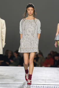 Model walks the runway in a light gray shift dress heavily embellished with silver beads and embroidery, with sleeves pushed up, paired with burgundy socks and red lace-up shoes.