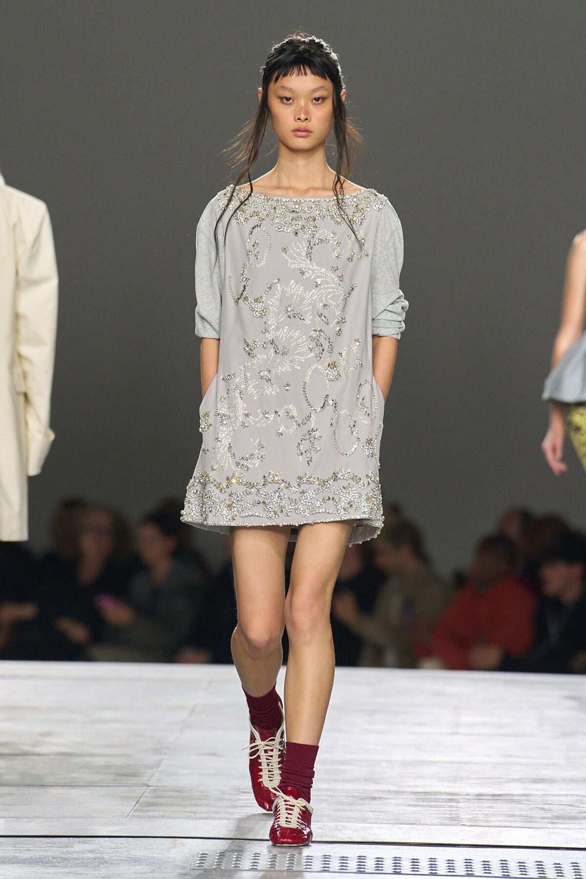 Model walks the runway in a light gray shift dress heavily embellished with silver beads and embroidery, with sleeves pushed up, paired with burgundy socks and red lace-up shoes.