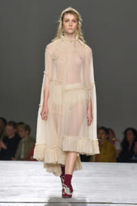 Model on a runway wearing a sheer cream dress with ruffled edges and long sleeves, paired with burgundy socks and lace-up shoes.