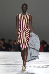 Model walking a fashion runway in a burgundy and beige striped knee-length dress with a belt and chunky necklace, holding a gray coat and patterned sneakers.