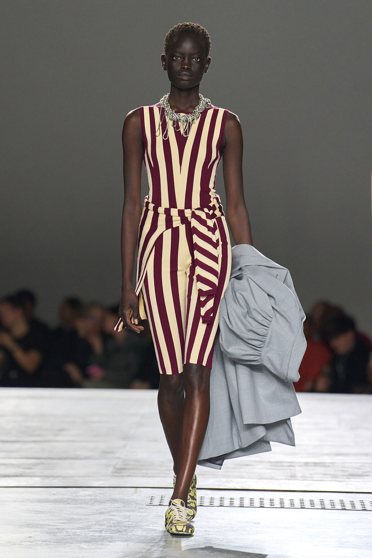 Model walking a fashion runway in a burgundy and beige striped knee-length dress with a belt and chunky necklace, holding a gray coat and patterned sneakers.