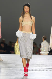 Model walking the runway in a beige lace bralette, a gray embroidered midi skirt, oversized white ruffled cuffs, and red socks with red lace-up shoes.