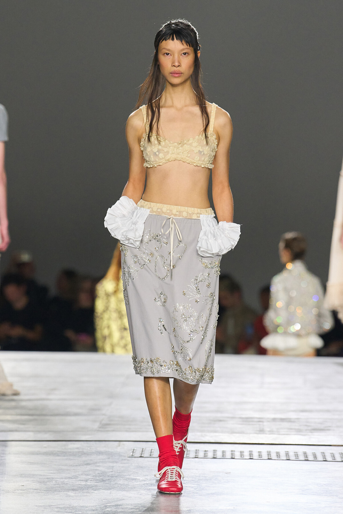 Model walking the runway in a beige lace bralette, a gray embroidered midi skirt, oversized white ruffled cuffs, and red socks with red lace-up shoes.