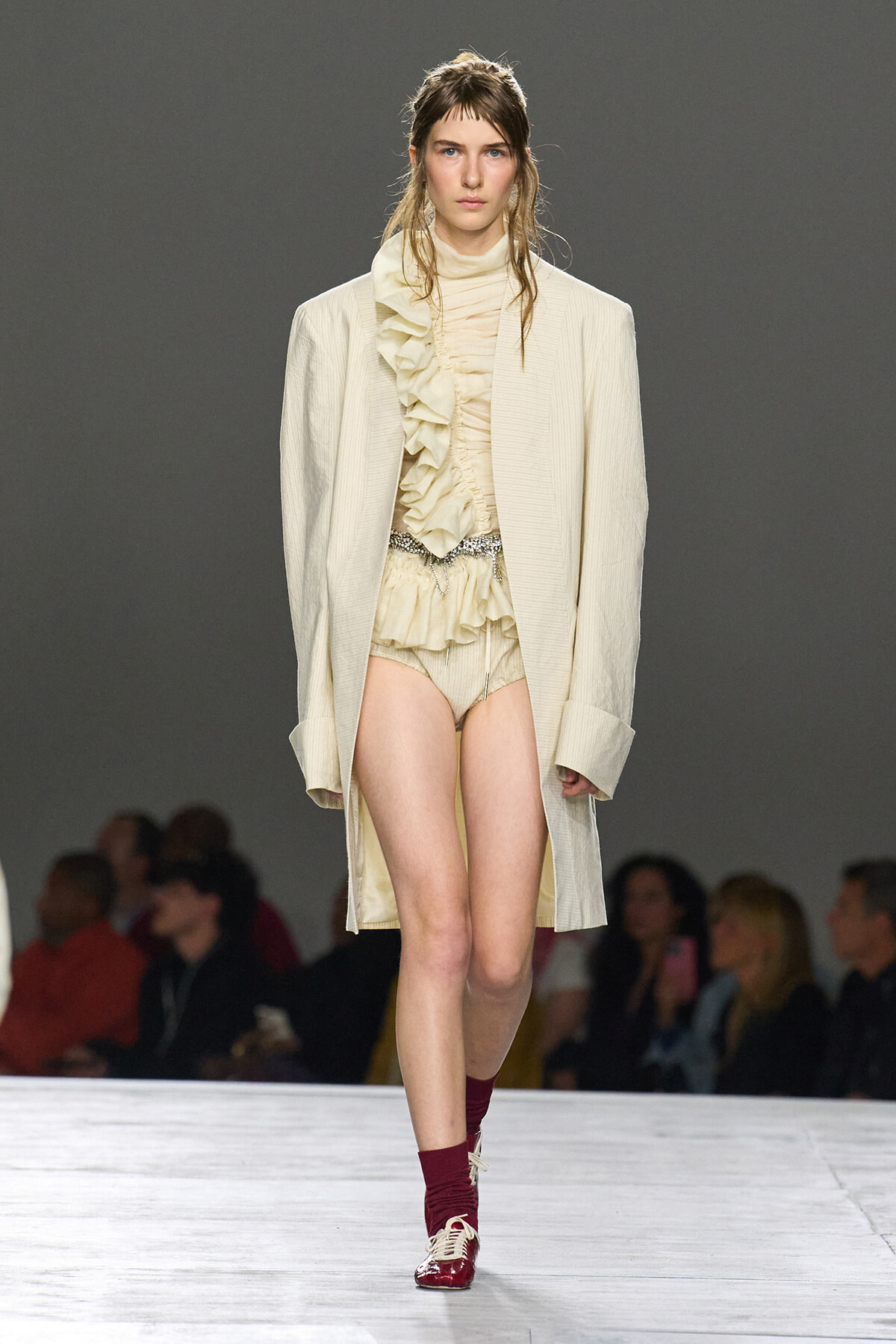 Model walking the runway in a cream ruffled blouse, matching shorts, and an oversized beige coat with burgundy socks and shiny oxford shoes.