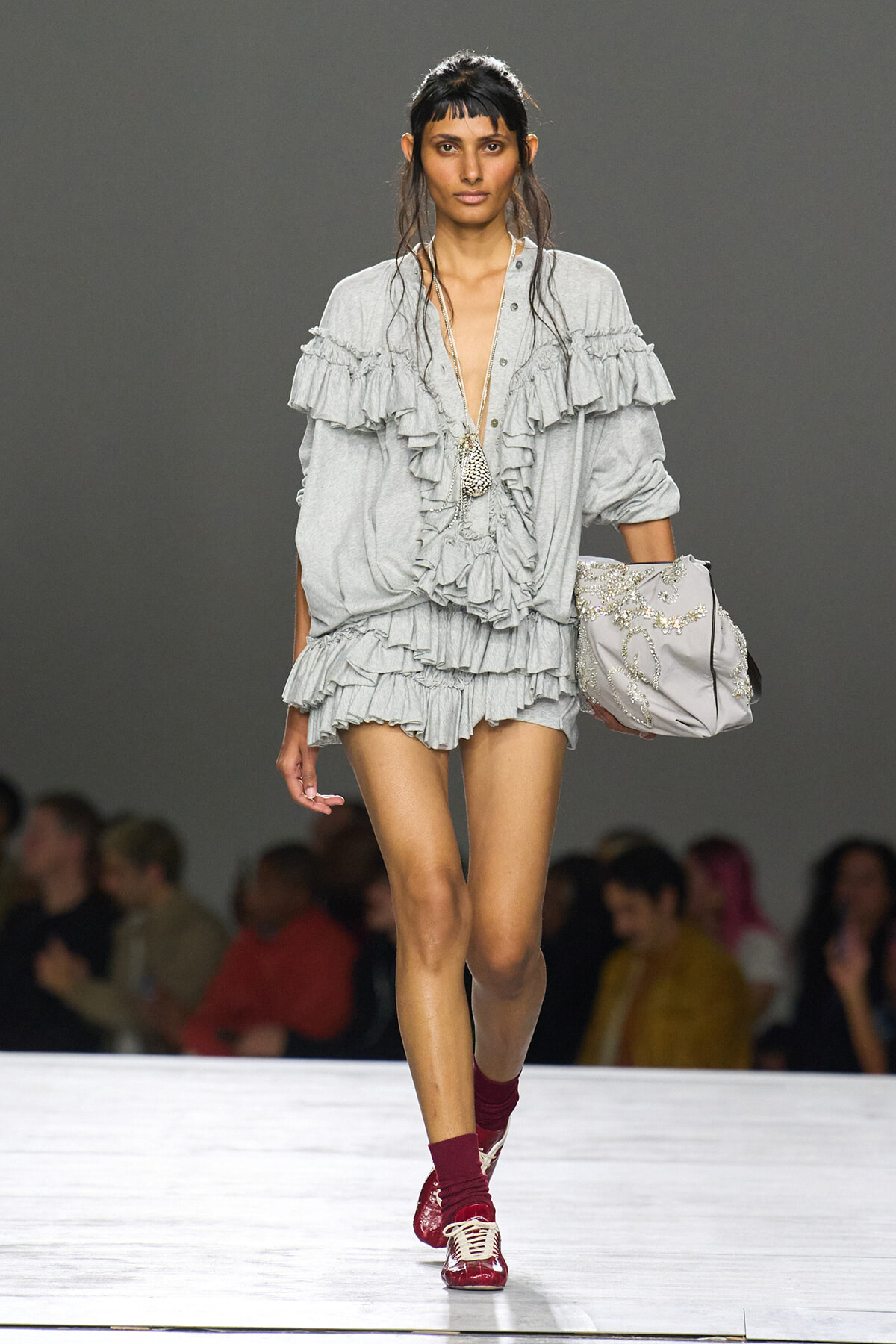 Model walking a runway in a light gray, ruffled romper with layered sleeves and shorts, carrying a beaded embroidered bag.