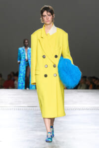 Model on the runway wearing a bright yellow, knee-length double-breasted coat with large buttons, holding a blue fluffy clutch, blue-pattern sneakers, audience in the background.