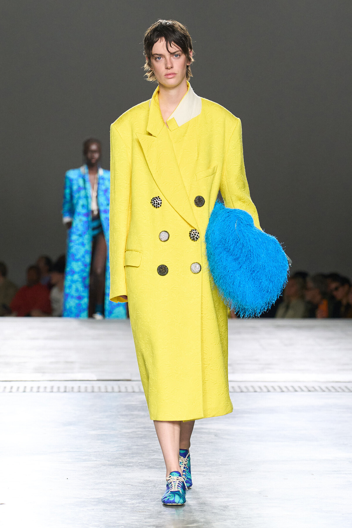 Model on the runway wearing a bright yellow, knee-length double-breasted coat with large buttons, holding a blue fluffy clutch, blue-pattern sneakers, audience in the background.