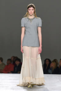 Model on a runway wearing a light gray short-sleeve top with a statement jeweled necklace and a cream, tiered sheer skirt.