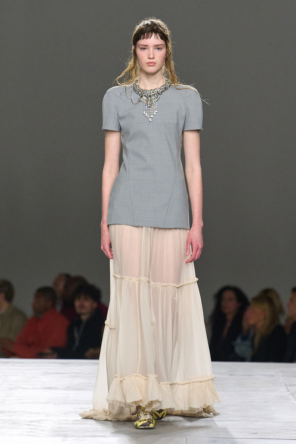 Model on a runway wearing a light gray short-sleeve top with a statement jeweled necklace and a cream, tiered sheer skirt.