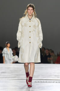 Model walking the runway in a long ivory coat with cream floral embroidery and gold buttons, paired with burgundy socks and red lace-up shoes.
