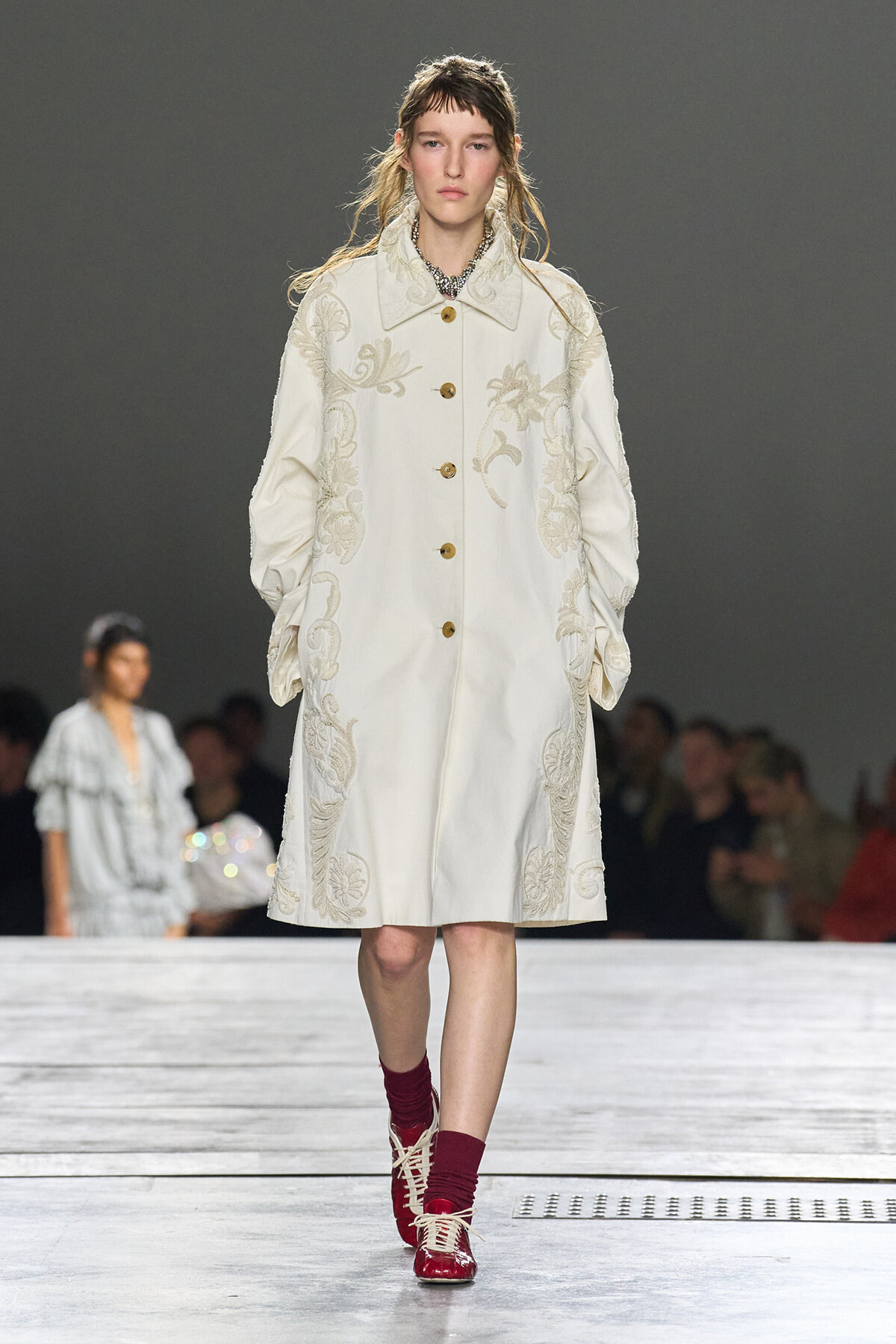 Model walking the runway in a long ivory coat with cream floral embroidery and gold buttons, paired with burgundy socks and red lace-up shoes.