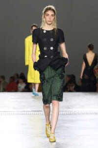 Model walking a runway in a black peplum top with silver circular embellishments and green embroidered knee-length shorts, yellow lace-up shoes present.