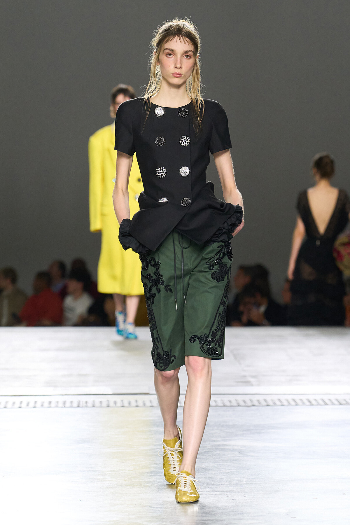 Model walking a runway in a black peplum top with silver circular embellishments and green embroidered knee-length shorts, yellow lace-up shoes present.