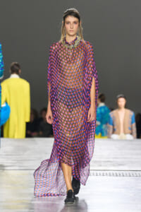 Model walks the runway in a sheer, polka-dot purple gown with a beaded collar and black sneakers while others in colorful outfits watch backstage.