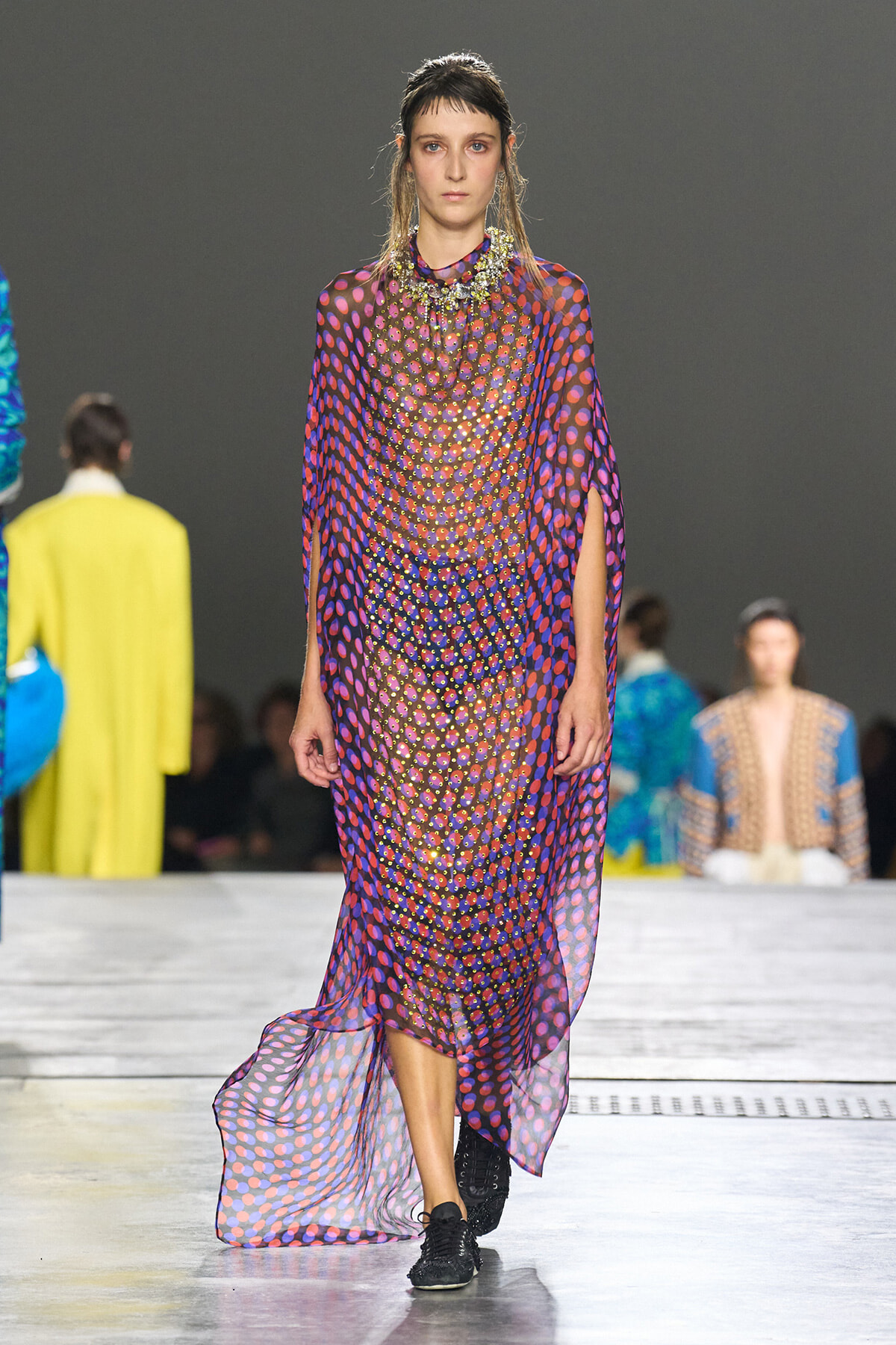 Model walks the runway in a sheer, polka-dot purple gown with a beaded collar and black sneakers while others in colorful outfits watch backstage.