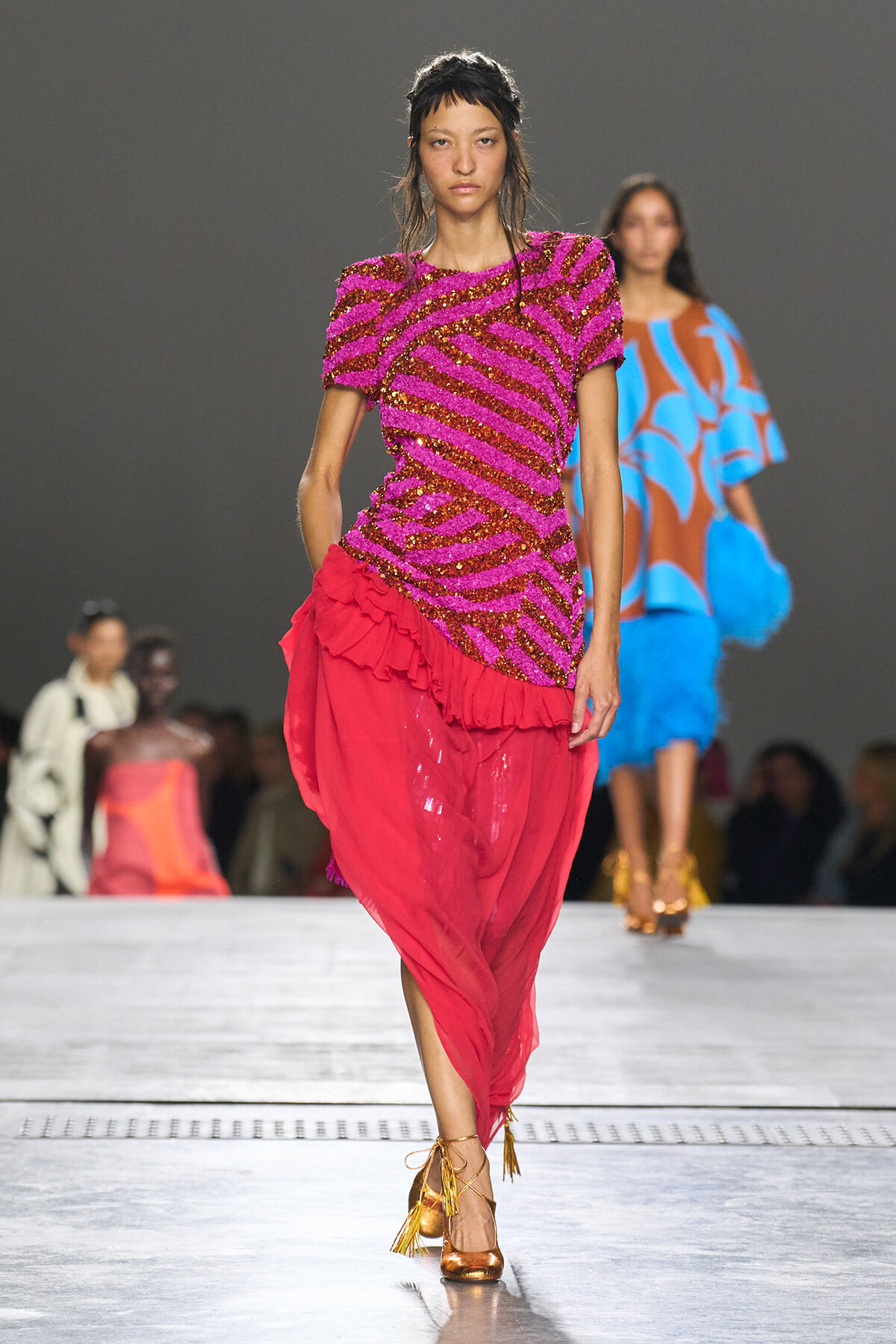 Model walking the runway in a vibrant pink sequined top with bold diagonal stripes and a voluminous red skirt with ruffles; gold sandals complete the look.