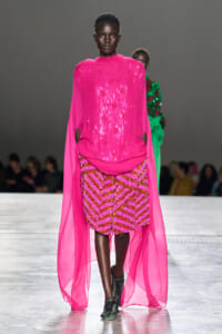 Model walking the runway in a bright pink sheer cape over a pink and gold sequin-patterned outfit.