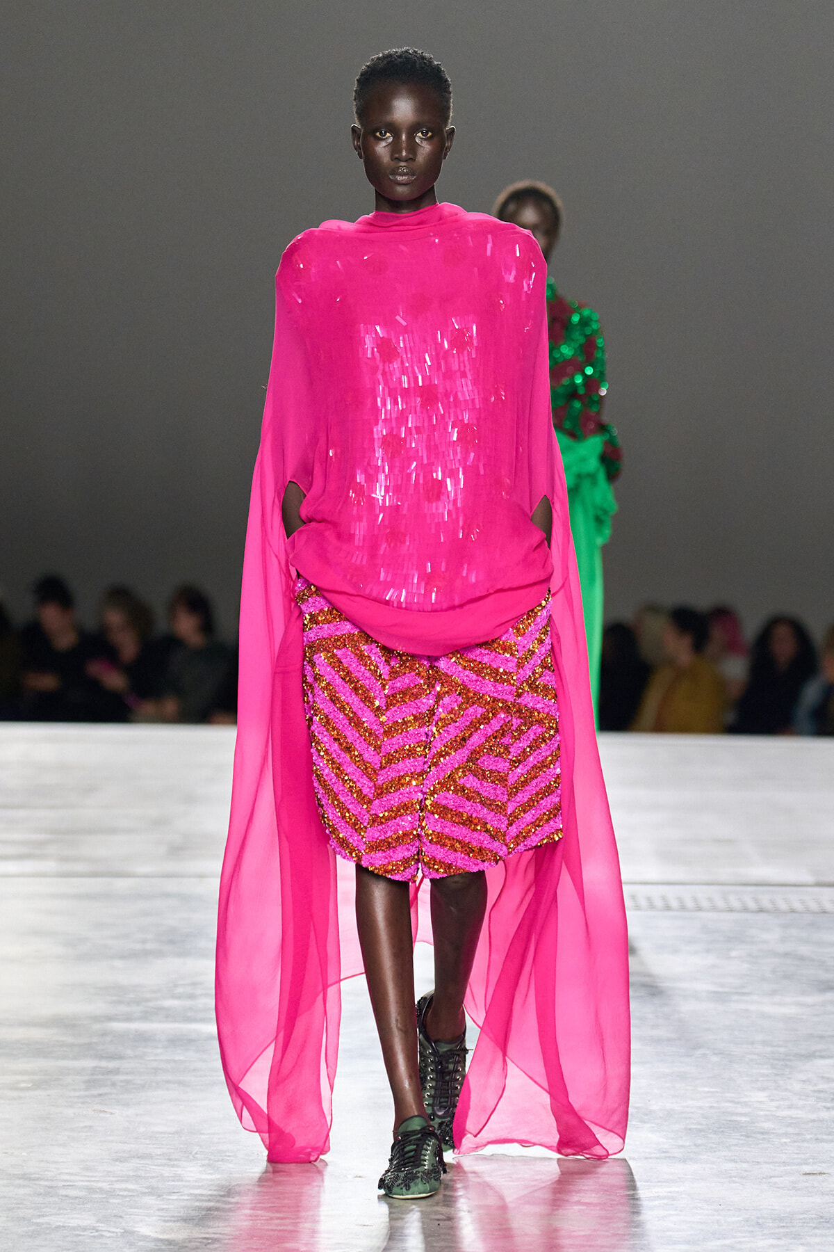Model walking the runway in a bright pink sheer cape over a pink and gold sequin-patterned outfit.