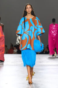 Model walking a runway in an oversized blue-brown abstract print top with a bright blue shaggy skirt and matching clutch, gold tassel heels visible.