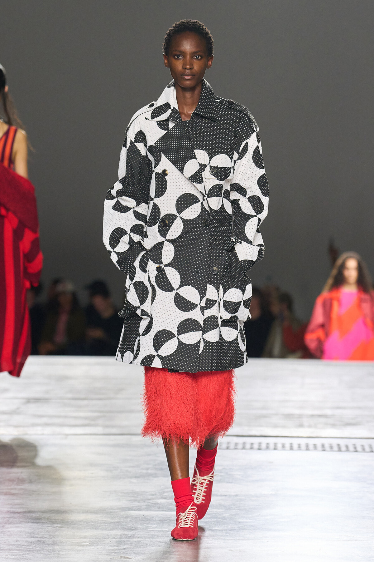 Model walking a runway in a loose black-and-white circle-patterned coat over a bright red feathered skirt, with red socks and red lace-up shoes.