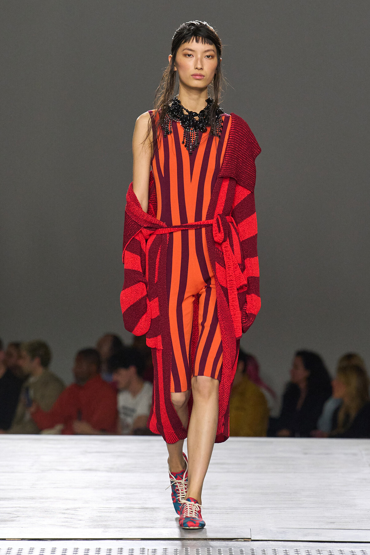 Model walking a fashion runway in a bold orange and red striped jumpsuit with a chunky black beaded necklace and a red striped cardigan draped over her shoulders.