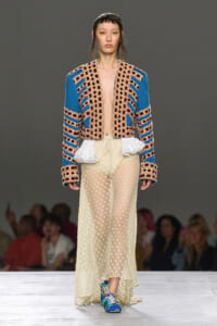 Runway model wearing a blue and cream embroidered cropped jacket with geometric patterns, a sheer polka-dot skirt with white ruffles, and blue sneakers.