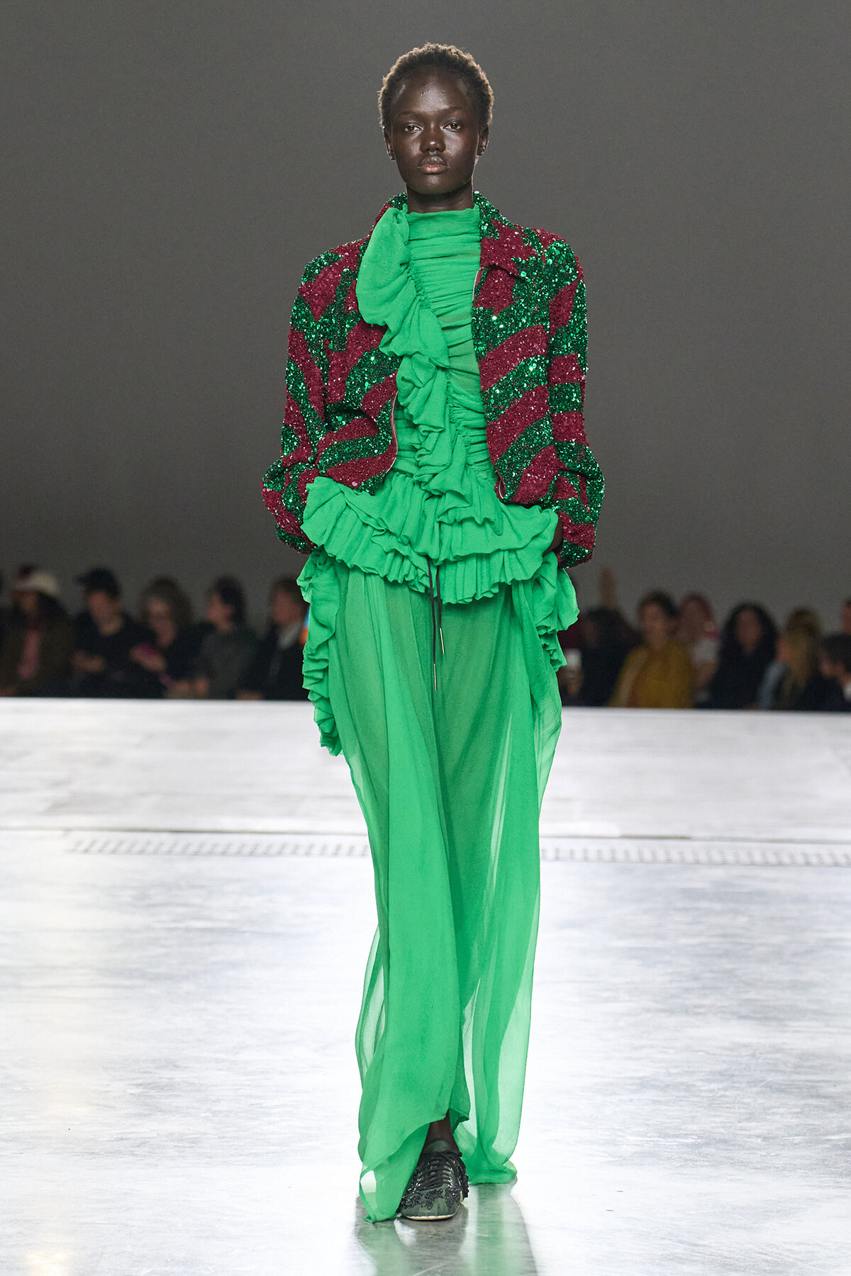 Model walking a runway in a vivid green, layered, ruffled gown paired with a red-and-green sequined jacket over it.