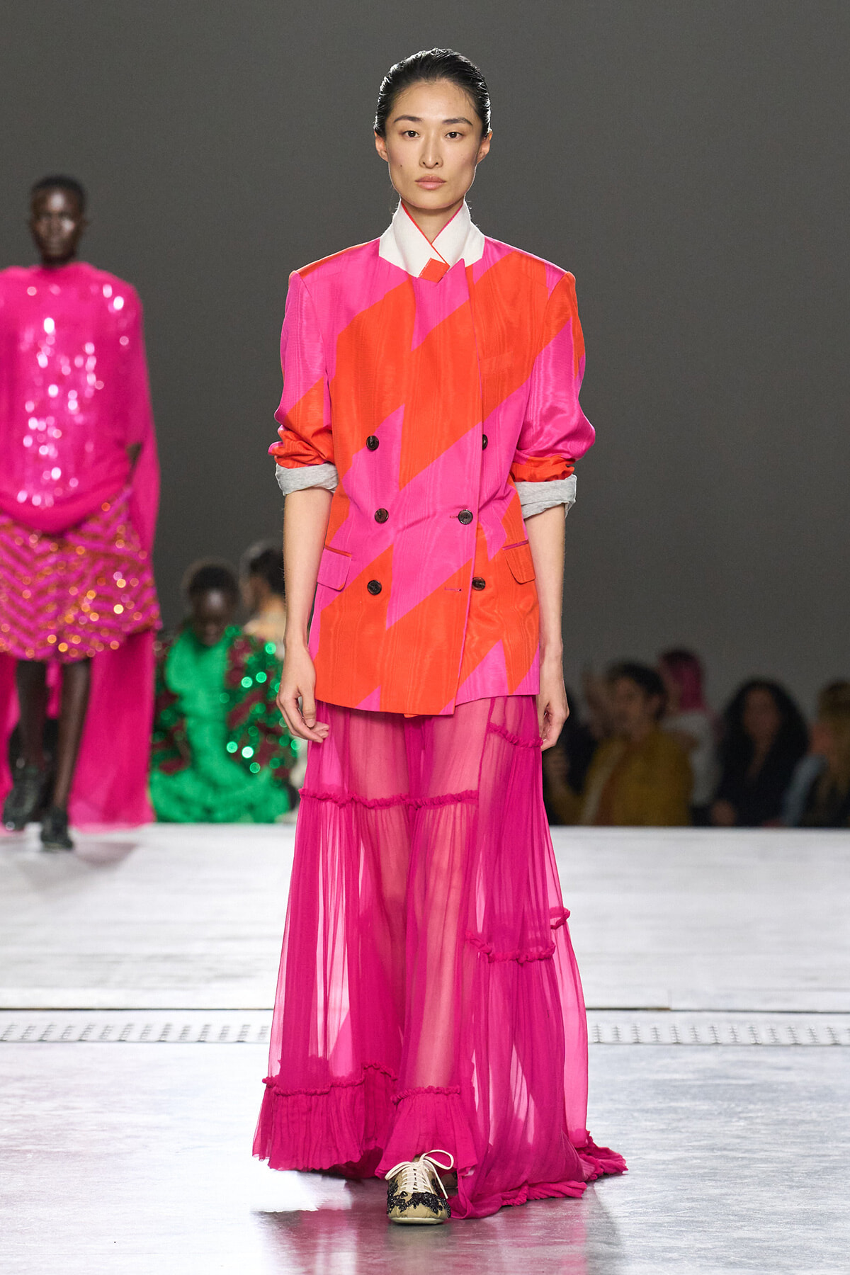 Model on runway wearing a pink-orange geometric blazer with rolled sleeves and a sheer magenta tiered skirt.