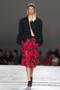 Model walking a runway in a black textured jacket over a bright pink floral embroidered midi skirt, with black shoes.