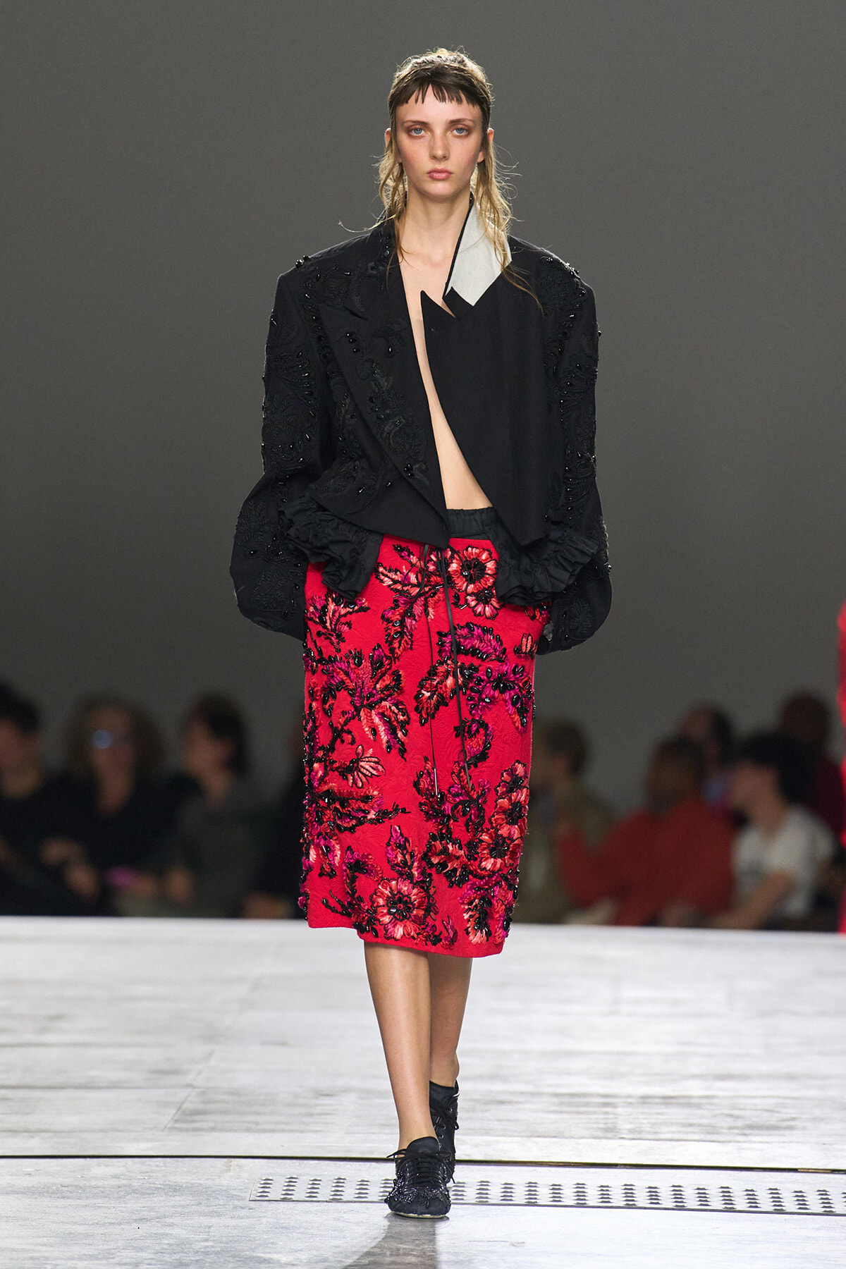 Model walking a runway in a black textured jacket over a bright pink floral embroidered midi skirt, with black shoes.