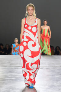 Model walks the runway in a loose red and white abstract print maxi dress; audience blurred in the background.