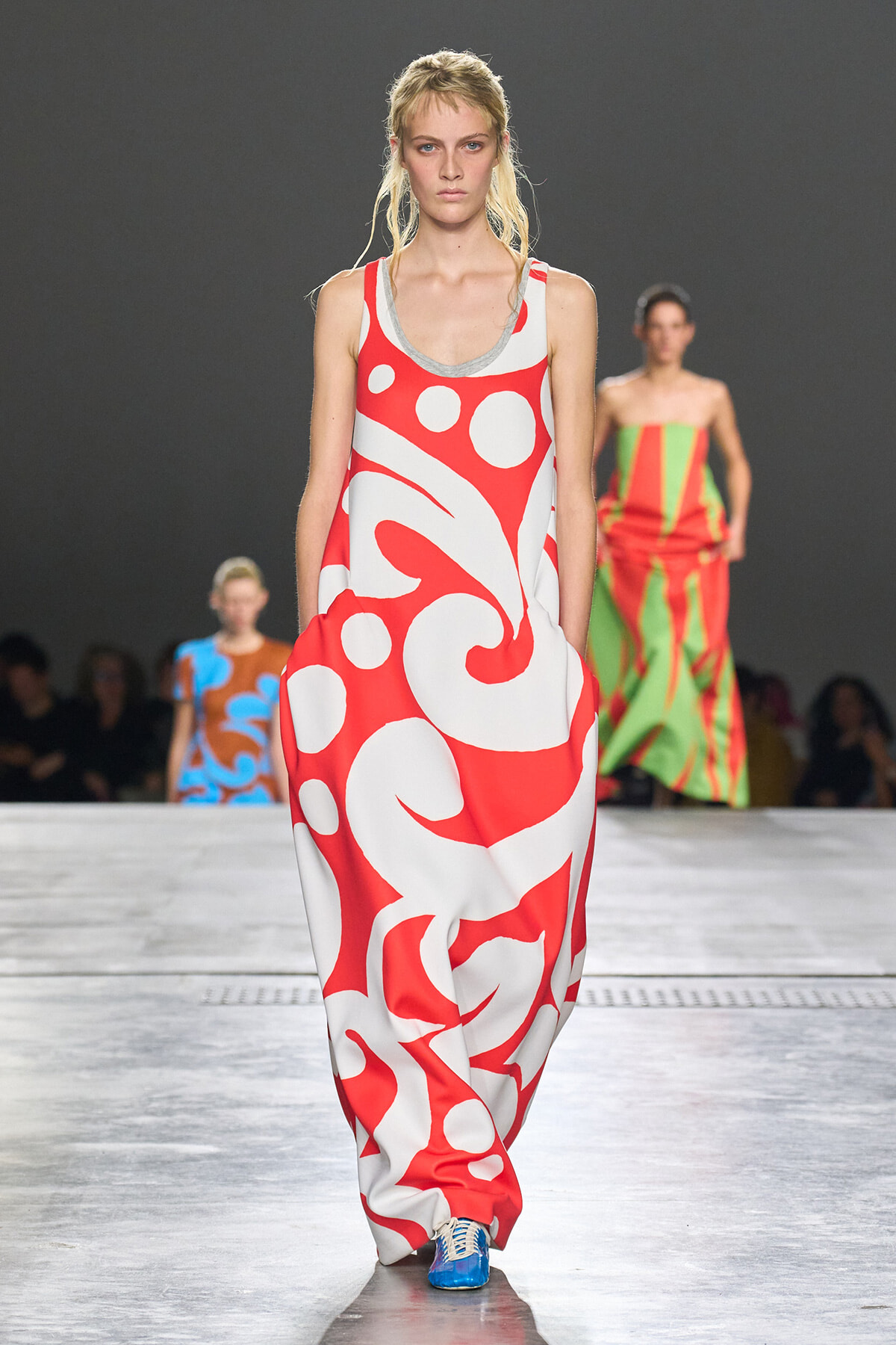 Model walks the runway in a loose red and white abstract print maxi dress; audience blurred in the background.