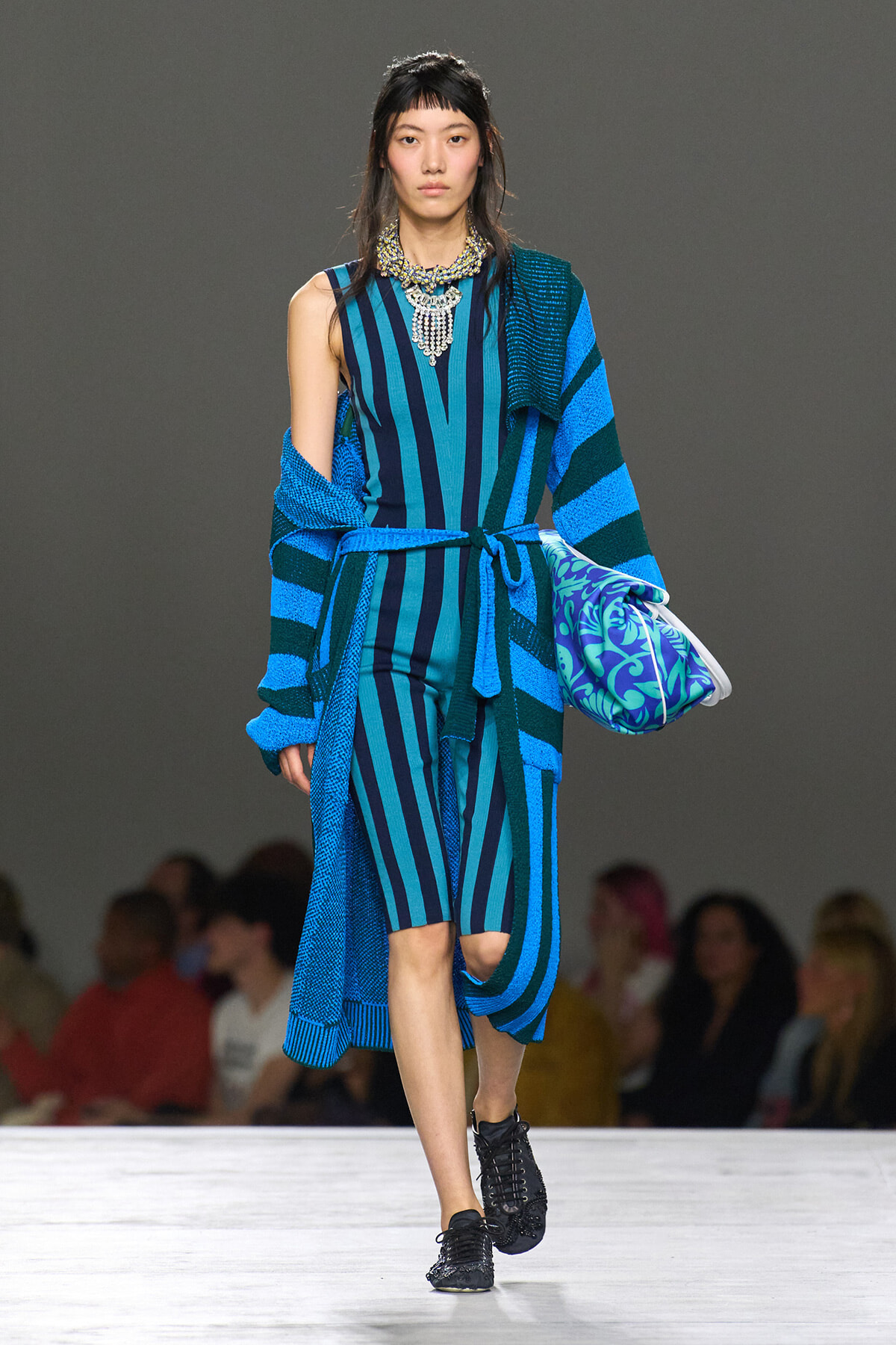 Model walking a runway in a blue striped jumpsuit with a matching belt, paired with a striped cardigan draped over the shoulders and a blue patterned bag; bold necklace and black sneakers visible.